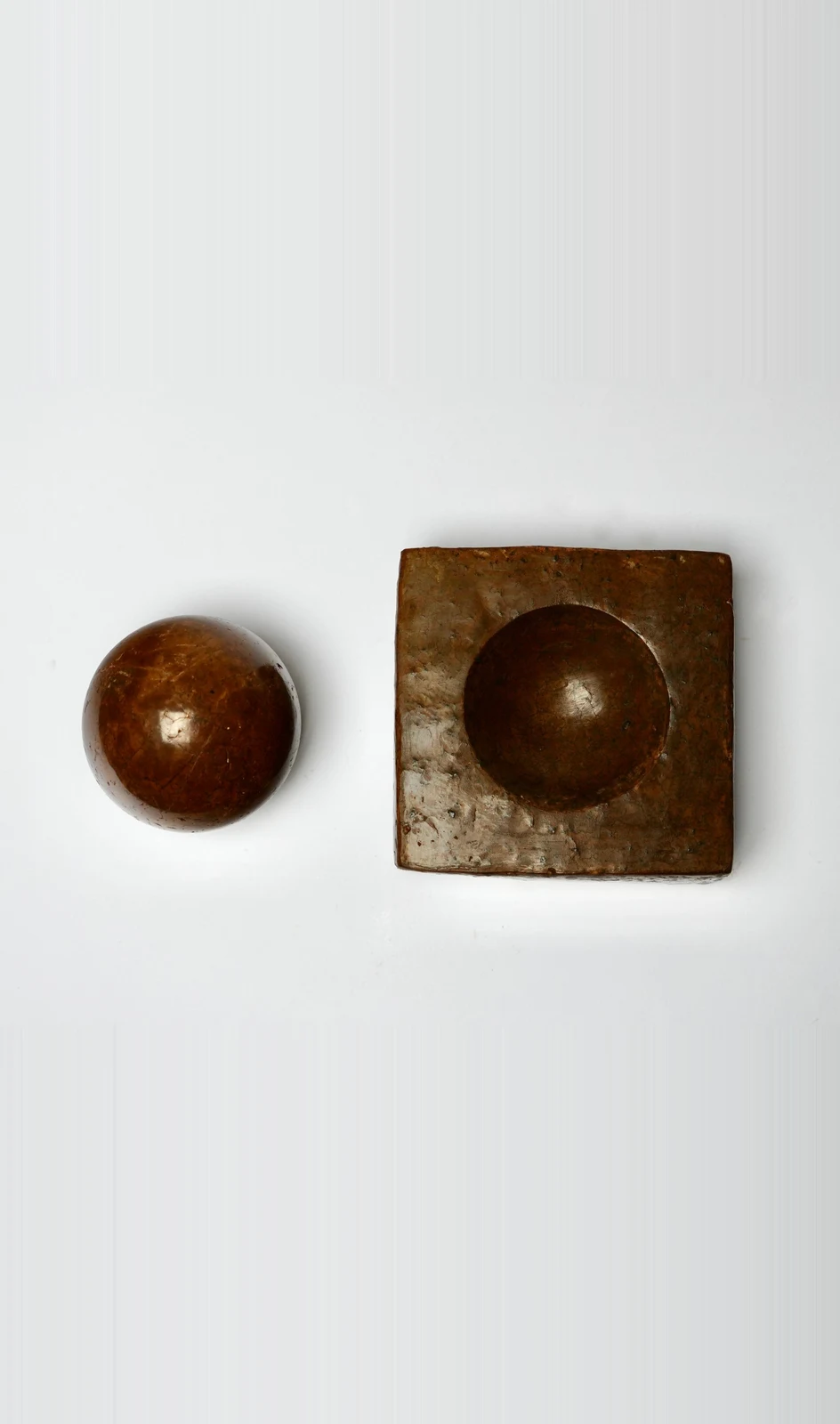 Dorodango Pair II by Edie Evans, a polished clay sphere beside a square block with a round hollow, seen from above