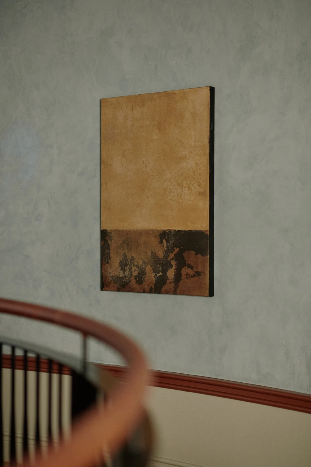 A painting by Alexandra Yan Wong with an ochre field above a dark band, hung on a blue plaster wall
